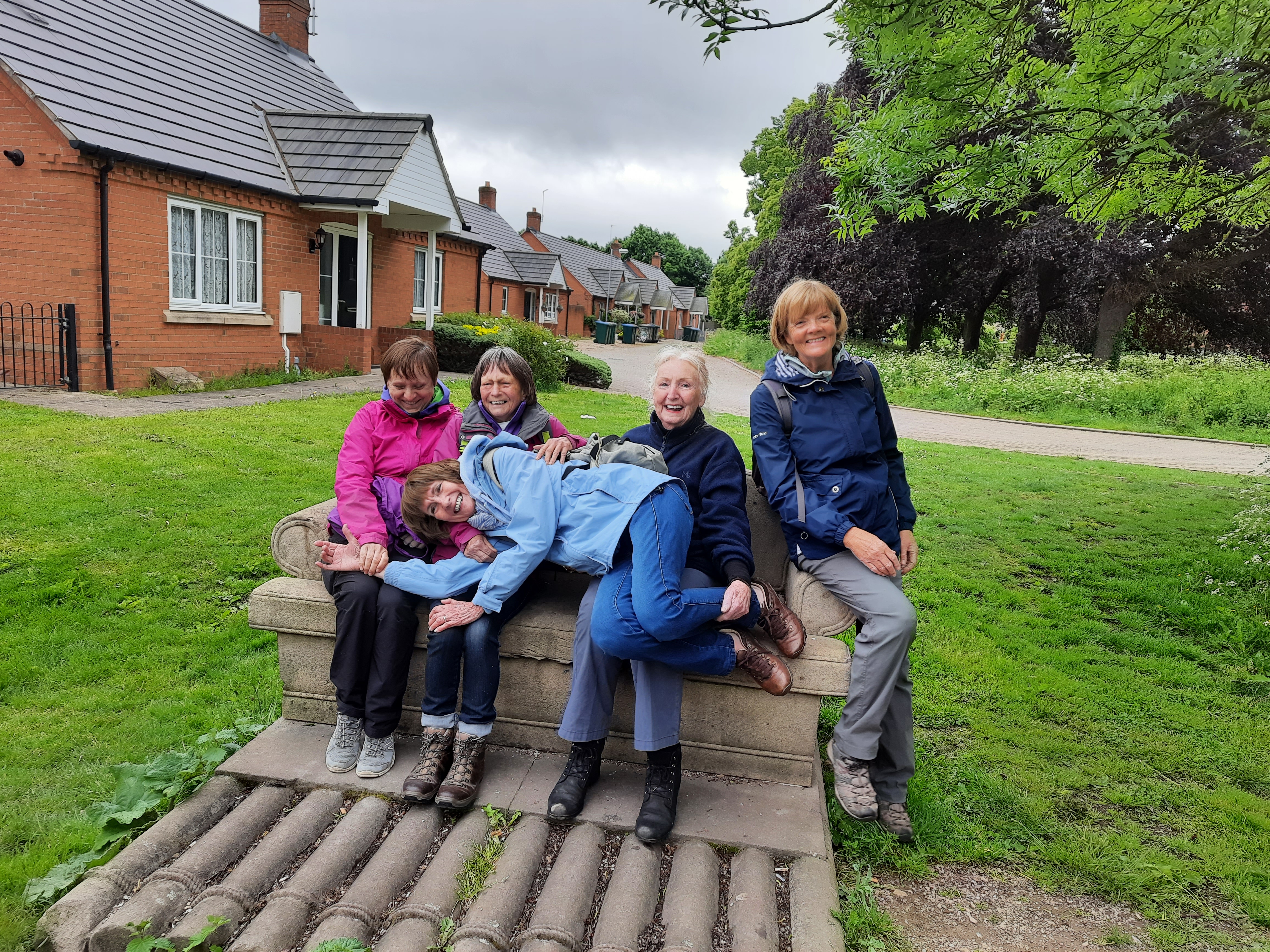 4 women sit on a bench with another lying across their legs.  All are laughing. 