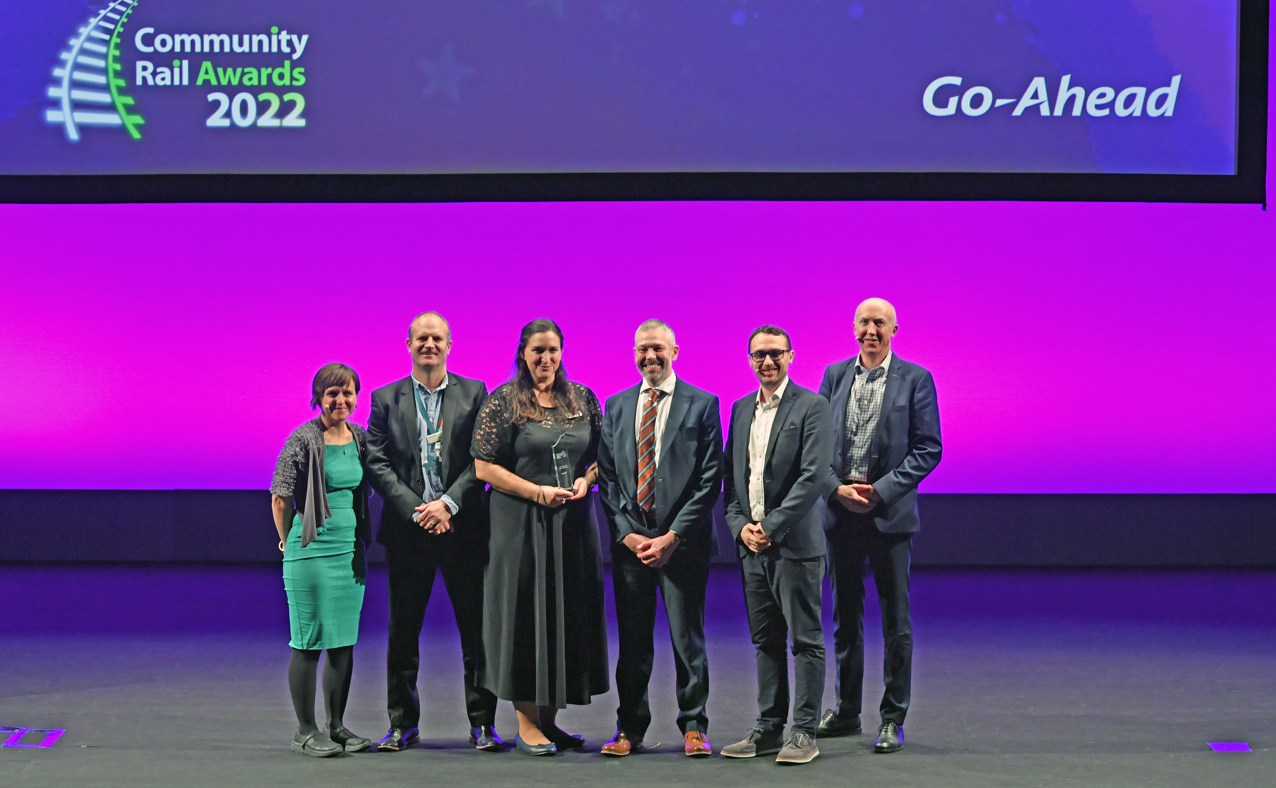 6 people, 4 men and 2 women, stand in front of a pinky purple screen with one of the women holding an award